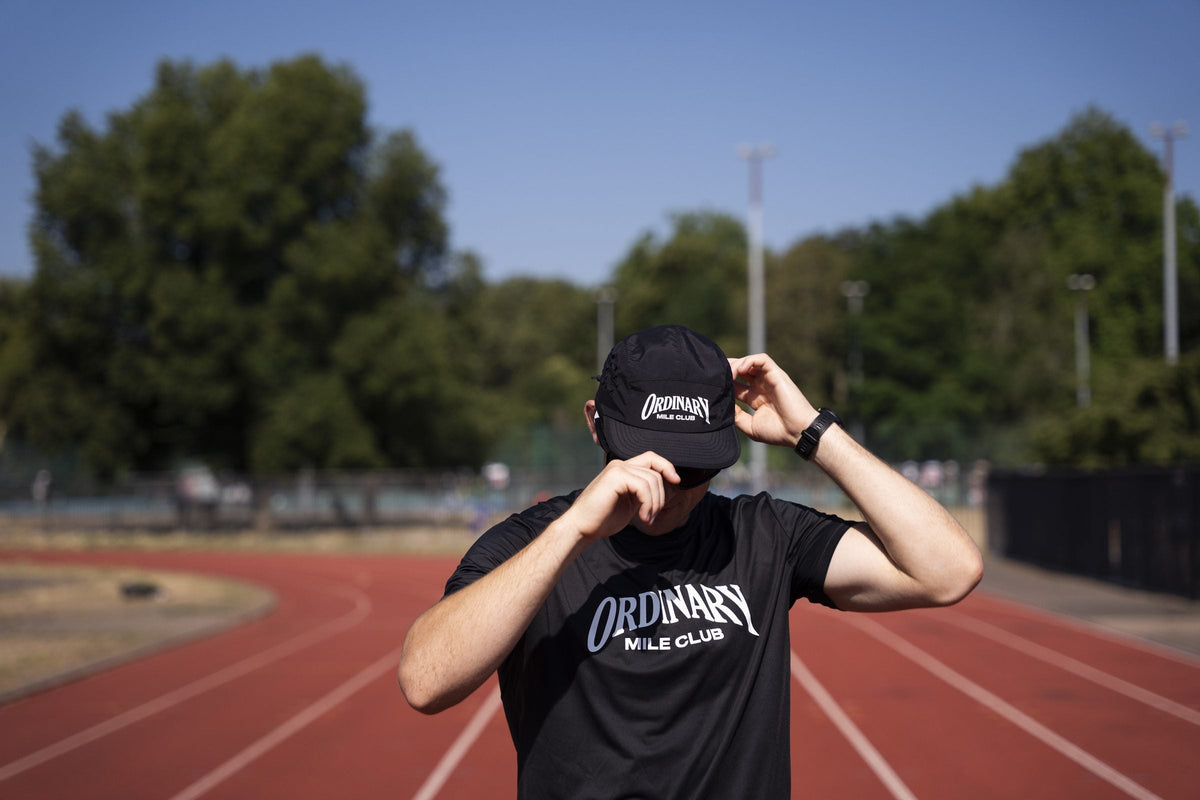 Black Ordinary Mile Club running cap laid flat showing the curved peak, mesh panel and technical sweatband