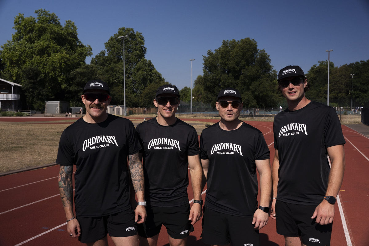 Four Ordinary Mile Club runners in black kit standing on a flat running track, ready for a fast parkrun PB attempt